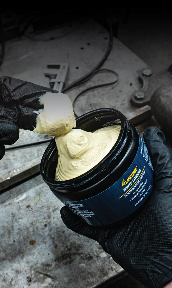 A person wearing black gloves is scooping white lithium grease from a blue container with a label in a workshop setting.