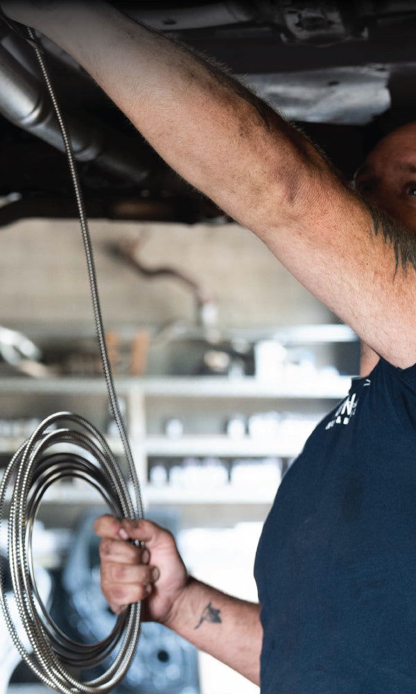 Mechanic in a garage holds a coiled metal hose while working under a car. The background shows blurred shelves with tools and parts, conveying focus and expertise.